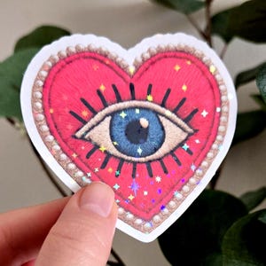 Peut inclure: Autocollant en forme de cœur avec un motif brodé rouge représentant un œil bleu au centre. L'œil est entouré de cils noirs et d'éclats d'étoiles. L'autocollant est bordé d'une bordure perlée.
