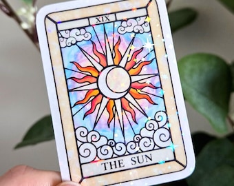 Puede incluir: Una pegatina rectangular con el diseño de la carta del tarot del Sol. La tarjeta tiene un borde color crema y presenta un sol con una luna creciente en el centro, rodeado de rayos de luz. El fondo es una mezcla de azul, rosa y naranja.