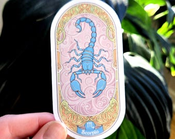 Könnte beinhalten: Ein ovaler Aufkleber mit einer hellblauen Skorpion-Illustration, dem Sternzeichen Skorpion. Das Design ist vor einem rosa Hintergrund mit Wirbelmustern platziert, umrahmt von verzierten goldenen und cremefarbenen Rändern. Das Wort "Scorpio" steht auf einem blauen Banner.