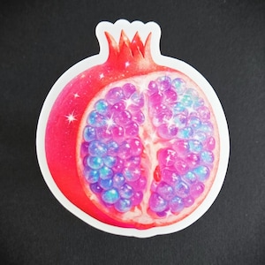 May include: A sticker featuring a cut pomegranate with a red rind and crown. The inside is filled with pink and blue jewel-toned seeds, with sparkling highlights. The sticker has a white border.