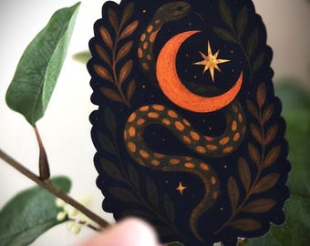 Può includere: Un adesivo con un design mistico raffigurante un serpente, una luna crescente e una stella. Il serpente è marrone e verde, avvolto attorno a una luna crescente arancione e una stella dorata, su uno sfondo blu scuro. Il design è incorniciato da foglie marroni.