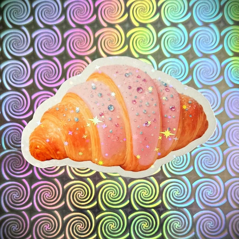 May include: A sticker featuring a croissant with pink icing and sparkling decorations. The pastry is orange-brown with a white border. The background is a holographic pattern of swirling rainbow colours.
