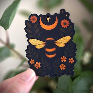 May include: A sticker featuring a bee with orange and yellow wings and stripes, set against a dark blue background. The design includes an orange crescent moon, star, and flowers, surrounded by green leaves.