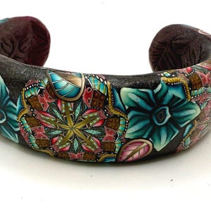 May include: A wide black bangle bracelet with a colorful floral design. The design features teal, pink, gold, and brown accents. The bracelet is made of polymer clay.