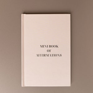 May include: A small, cream-coloured book with the title "MINI BOOK OF AFFIRMATIONS" in a simple font. The book is centred on a neutral grey background. The cover has a clean, minimalist design.