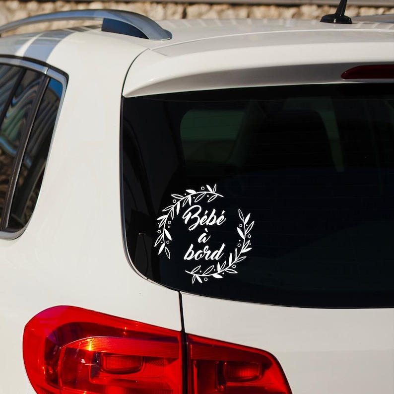 May include: White car with a white vinyl sticker on the back window. The sticker says "B&eacute;b&eacute; &agrave; bord" in a decorative font with a floral wreath around the text.