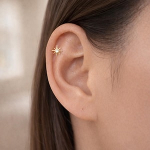 Op de afbeelding: Een goudkleurige oorpiercing in de vorm van een ster met een heldere edelsteen in het midden. Het sieraad is geplaatst in het bovenste kraakbeen van het oor en heeft een metallic afwerking en een stralende, veelpuntige stervorm.
