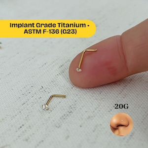 May include: Two gold-coloured, L-shaped nose rings, each with a small, clear gemstone. The text "Implant Grade Titanium - ASTM F-136 (G23)" is in a yellow box. The text "20G" is also visible.
