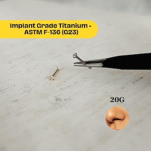 Può includere: Un piercing al naso color oro con una gemma trasparente, accanto a un paio di pinzette che tengono un perno simile. L'immagine include il testo "Implant Grade Titanium - ASTM F-136 (G23)" e "20G".