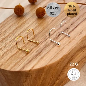 May include: Two pairs of nose rings on a wooden surface. One pair is silver and the other is gold plated. The silver pair is labeled "Silver 925" and the gold plated pair is labeled "18K Gold plated".