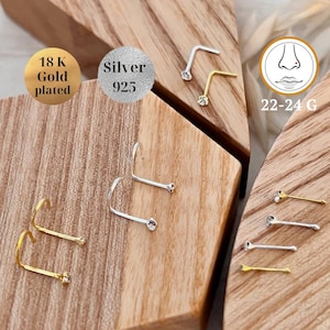 Op de afbeelding: Een close-up van een verscheidenheid aan neusringen met heldere strass steentjes. De ringen zijn gemaakt van zilver, verguld en roestvrij staal. De ringen worden gepresenteerd op een houten oppervlak. 22-24 G.