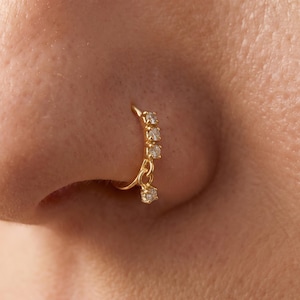 May include: A gold nose hoop featuring three small, clear, round-cut stones set along the upper curve and a single dangling clear stone charm attached by a small jump ring, creating a delicate and shimmering appearance on the nostril.
