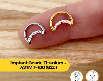 Könnte beinhalten: Zwei halbmondförmige Nasenringe, einer silberfarben und einer goldfarben, jeweils mit einer Reihe kleiner, klarer Steine. Die Ringe werden auf einem Finger vor einem Holzgrund gezeigt. Text auf einem gelben Banner: "Implant Grade Titanium - ASTM F-136 (G23)".