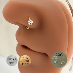 May include: A gold-plated nose ring featuring a flower design with clear gemstones. The ring is shown on a person's nose. The image also includes text indicating the ring is 18K gold plated and silver 925.