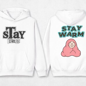 Peut inclure: Sweat à capuche blanc avec poche avant et capuche. Le devant affiche "Stay TRUE" dans une police usée. Le dos présente "STAY WARM" au-dessus d'un personnage de dessin animé enveloppé dans une couverture rose.