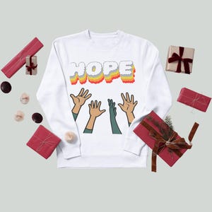Può includere: Felpa bianca con la scritta "HOPE" in un design colorato e stratificato. Sotto la parola ci sono illustrazioni di mani che si alzano. L'immagine è circondata da regali confezionati in toni rossi e marroni.