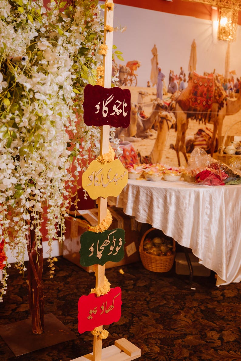 May include: A wooden signpost with four colorful, decorative signs in Urdu script. The signs are in maroon, yellow, green, and red. The background features a desert scene with camels and a table with food.