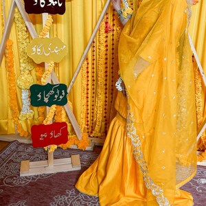 May include: A woman wearing a bright yellow traditional outfit with a matching scarf. The outfit has silver embroidery. The background features yellow fabric and decorative signs with text in a foreign language. Floral garlands and decorations are also present.