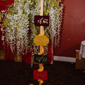 May include: A wooden signpost with four colorful, shaped signs in Urdu script. The signs are in maroon, yellow, green, and red. The signpost is decorated with yellow flowers and is set against a backdrop of white flowers and red fabric.