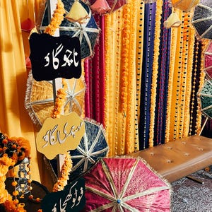 May include: A festive scene with colorful umbrellas, garlands, and signs. The umbrellas are in shades of red, blue, and gold, with intricate patterns. The signs have text in a foreign language. The background features draped fabric in yellow, purple, and blue.