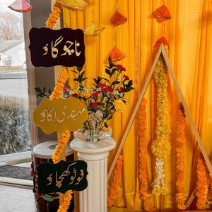 May include: A festive event setup with yellow and orange decorations. Includes hanging ornaments, floral garlands, and signs with Urdu text. A floral arrangement sits on a white pedestal. A wooden triangle frame is adorned with flowers.
