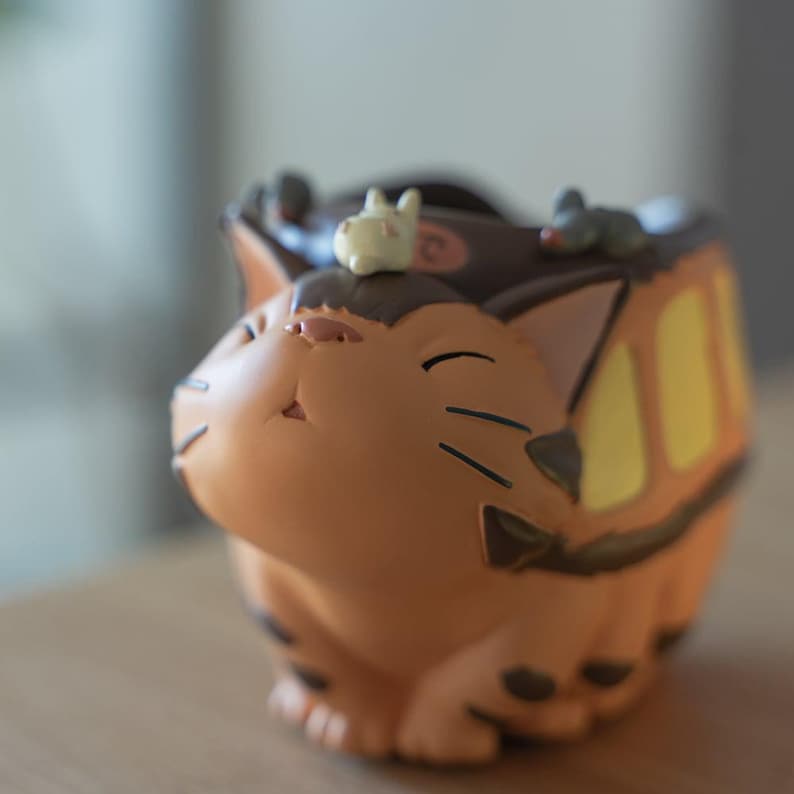 May include: A tan and brown cat-shaped planter with a small white bunny and gray mouse figurine on top. The planter has black stripes and yellow accents. The cat's face is detailed with black whiskers and a pink nose.