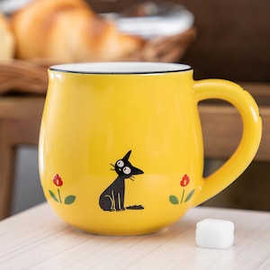 May include: A bright yellow ceramic mug with a rounded body and a curved handle. The mug features a black cat illustration centered on the front, flanked by two small red flowers with green stems and leaves. The rim of the mug is finished in black.