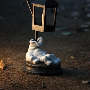 May include: A decorative lamp with a black lantern top and a white hand-shaped base. The lantern has a small candle inside. The base is a dark brown color and sits on a dirt surface. The lamp is a unique home decor item.