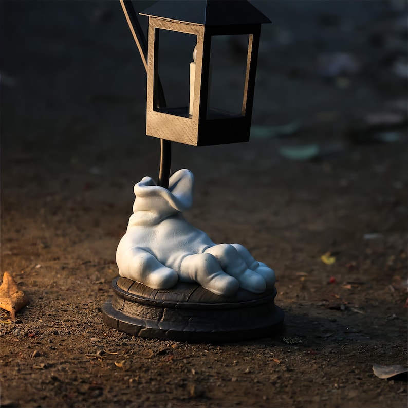 May include: A decorative lamp with a black lantern top and a white hand-shaped base. The lantern has a small candle inside. The base is a dark brown color and sits on a dirt surface. The lamp is a unique home decor item.