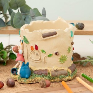 Puede incluir: Una maceta de cer&aacute;mica beige con dise&ntilde;o de tronco de &aacute;rbol, con un personaje de dibujos animados y un ni&ntilde;o trepando. La maceta est&aacute; decorada con hojas, bellotas y detalles coloridos. La maceta mide aproximadamente 13 cm de alto.