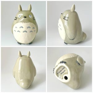 May include: A ceramic figurine of Totoro, a character from the animated film My Neighbor Totoro. The figurine is light gray with a cream-colored belly and gray markings. It has large eyes, small ears, and black whiskers. The bottom of the figurine has the text "Studio Ghibli Seloguchi".