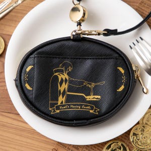 May include: Black oval coin purse with a gold zip and a lanyard. The purse features a gold illustration of a character from Howl's Moving Castle. The text "Howl's Moving Castle" is below the illustration. The purse is on a white plate.