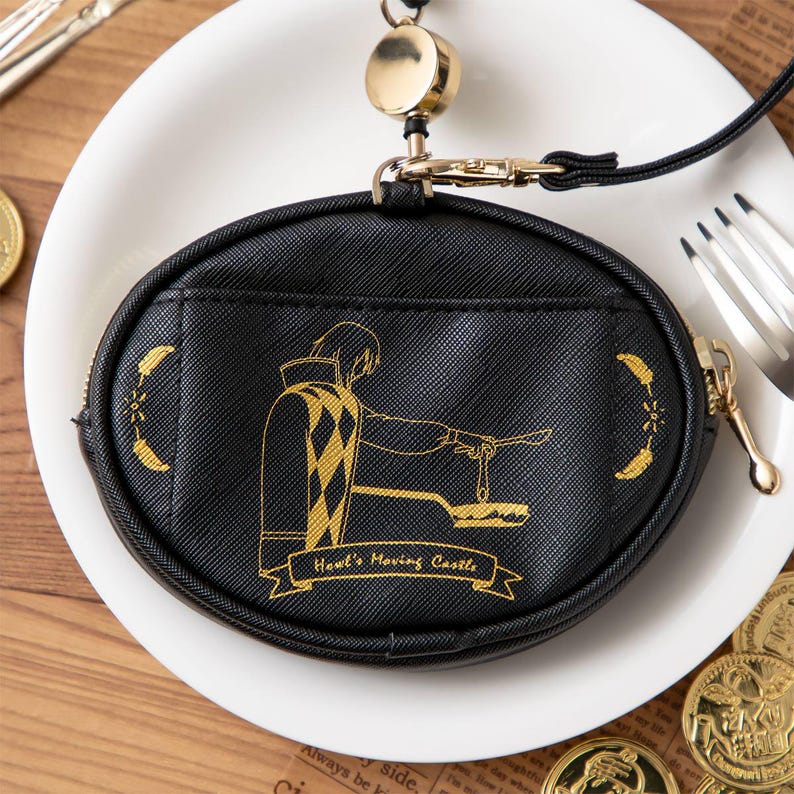 May include: Black oval coin purse with a gold zip and a lanyard. The purse features a gold illustration of a character from Howl's Moving Castle. The text "Howl's Moving Castle" is below the illustration. The purse is on a white plate.