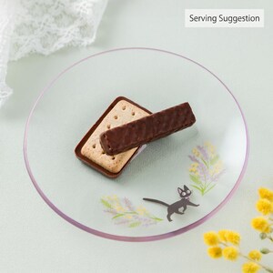 Op de afbeelding: Een rond, helder glazen bord met een kleine zwarte kat-illustratie en bloemaccenten in geel en paars. Een rechthoekig lichtgekleurd koekje met daarop een met pure chocolade omhulde wafelreep ligt in het midden. De tekst luidt Serving Suggestion.