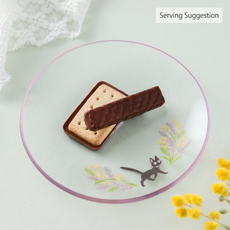 Op de afbeelding: Een rond, helder glazen bord met een kleine zwarte kat-illustratie en bloemaccenten in geel en paars. Een rechthoekig lichtgekleurd koekje met daarop een met pure chocolade omhulde wafelreep ligt in het midden. De tekst luidt Serving Suggestion.