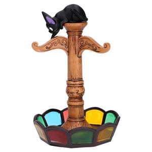 May include: A decorative incense holder featuring a black cat with purple accents perched atop a wooden pillar. The pillar has decorative carvings and supports a colorful, stained-glass style base with various colored panels.