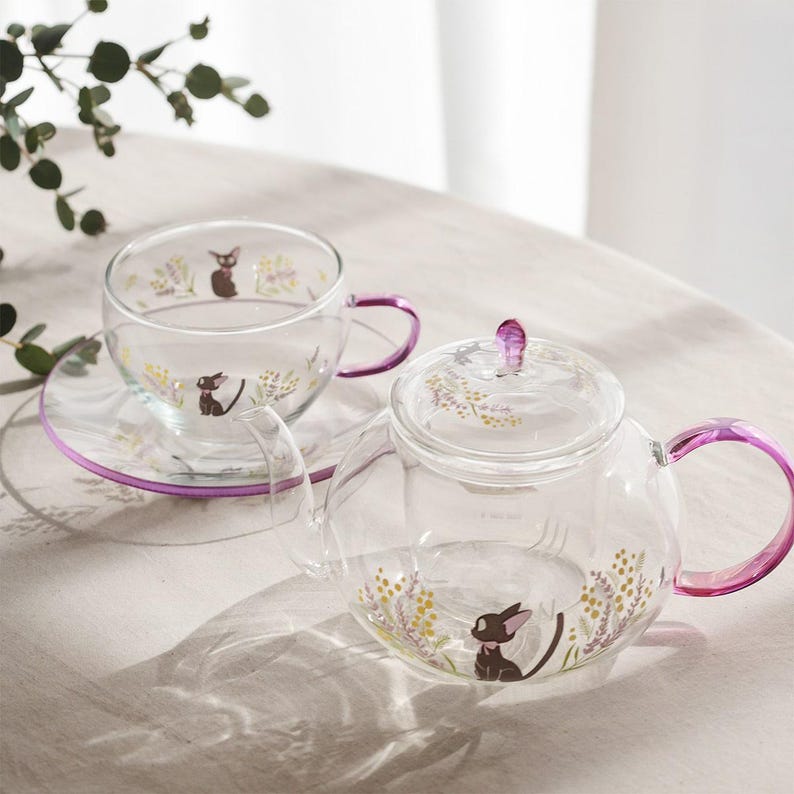 Puede incluir: Una tetera y una taza a juego con plato de cristal transparente, con asas y pomo de tapa de color morado. Las superficies de cristal est&aacute;n decoradas con ilustraciones de un peque&ntilde;o gato negro rodeado de ramilletes de flores amarillas y moradas.