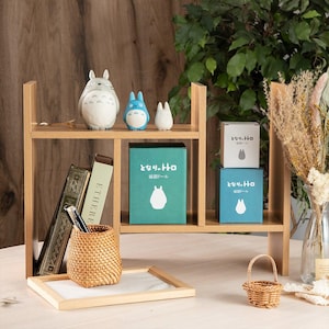 May include: A wooden shelf unit displays Totoro figurines, boxes with Japanese text, books, and a woven basket. The shelf is light brown, with a small white tray and a miniature woven basket on the bottom shelf.