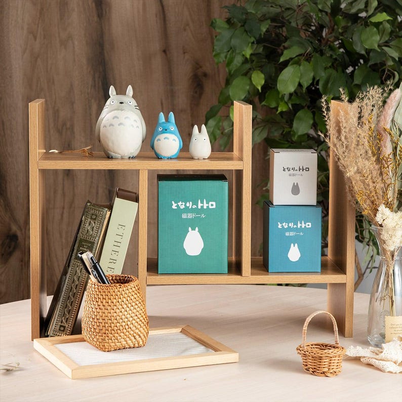 May include: A wooden shelf unit displays Totoro figurines, boxes with Japanese text, books, and a woven basket. The shelf is light brown, with a small white tray and a miniature woven basket on the bottom shelf.