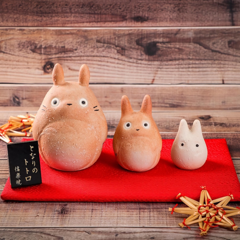 May include: Three ceramic Totoro figures in varying sizes, arranged on a red surface. The largest is light brown, the middle one is orange, and the smallest is white. A small black sign with Japanese text is in front of the largest figure.