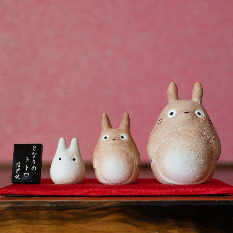 May include: Three ceramic Totoro figurines of varying sizes, arranged on a red mat against a pink background. The largest figure is beige with white eyes and whiskers. A small black sign with Japanese text is on the left.