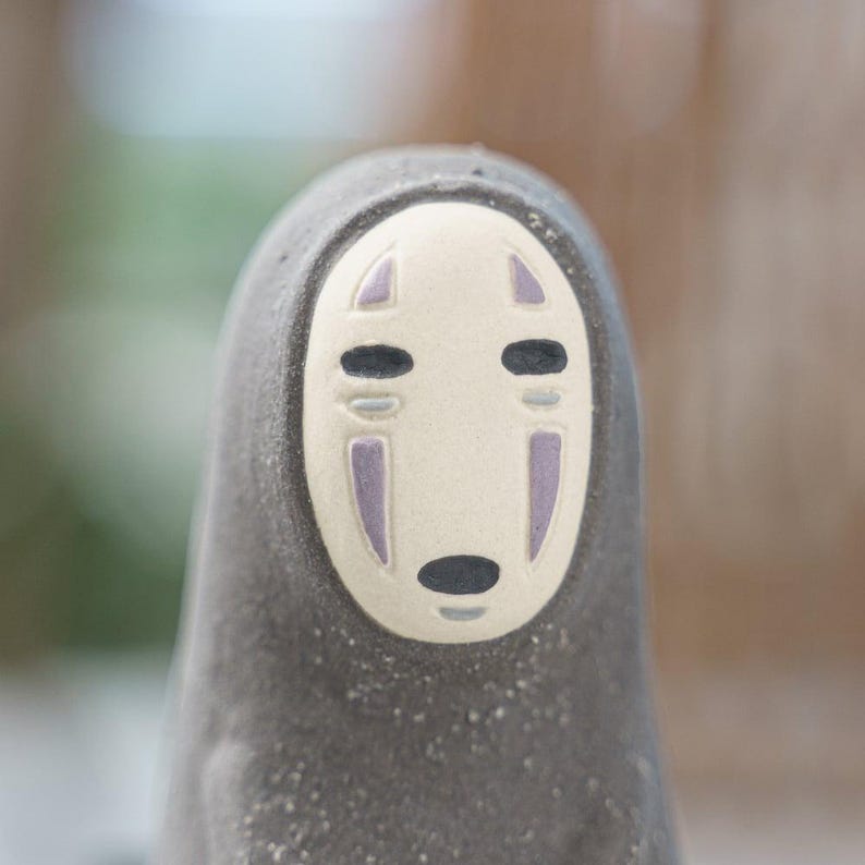 Puede incluir: Una figura de cer&aacute;mica con un rostro ovalado liso de color crema y marcas estilizadas en negro y morado. El cuerpo es de color gris oscuro, texturizado y redondeado, estrech&aacute;ndose hacia arriba, asemej&aacute;ndose a un personaje encapuchado.
