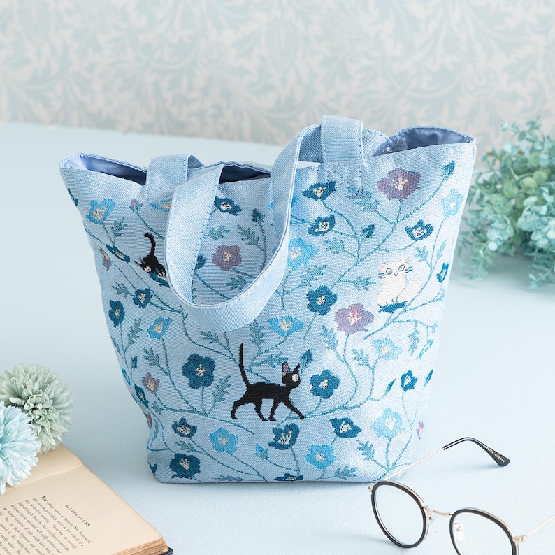 May include: A light blue tote bag with a floral pattern and embroidered black and white cats. The bag has two handles.