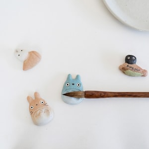 Puede incluir: Cuatro reposapalillos de cer&aacute;mica con figuras de animales estilizadas, incluyendo una criatura azul claro, una beige, una blanca y una figura de cabeza oscura con texto en japon&eacute;s, con un pincel de madera apoyado sobre la pieza azul.