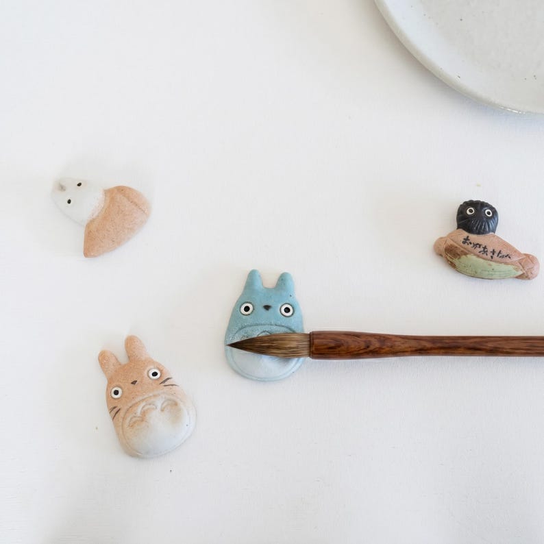 Puede incluir: Cuatro reposapalillos de cer&aacute;mica con figuras de animales estilizadas, incluyendo una criatura azul claro, una beige, una blanca y una figura de cabeza oscura con texto en japon&eacute;s, con un pincel de madera apoyado sobre la pieza azul.