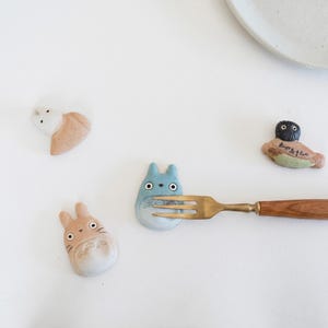 Puede incluir: Cuatro reposa palillos de cer&aacute;mica con forma de criaturas de dibujos animados, incluyendo uno azul, uno bronceado, uno blanco y uno de pelo oscuro con texto japon&eacute;s. Un tenedor de metal con mango de madera descansa sobre la figura azul.