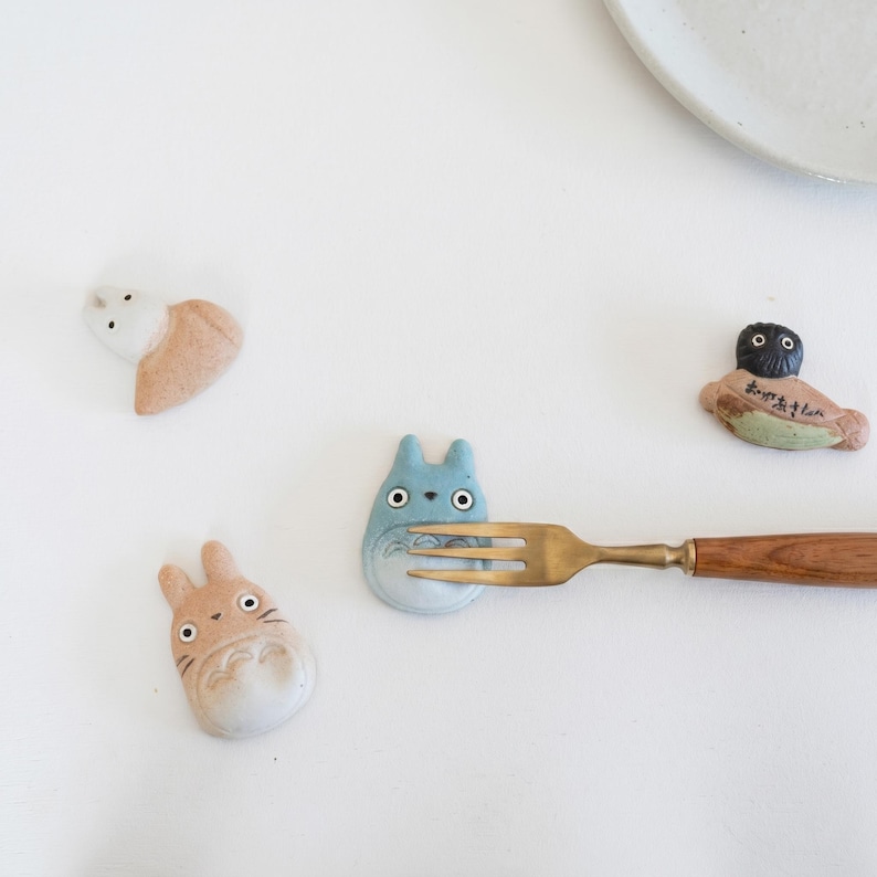 Puede incluir: Cuatro reposa palillos de cer&aacute;mica con forma de criaturas de dibujos animados, incluyendo uno azul, uno bronceado, uno blanco y uno de pelo oscuro con texto japon&eacute;s. Un tenedor de metal con mango de madera descansa sobre la figura azul.
