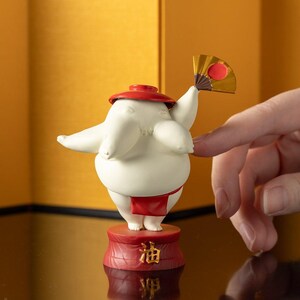 Peut inclure: Figurine blanche repr&eacute;sentant un lutteur de sumo avec un chapeau et un pagne rouges, tenant un petit &eacute;ventail. La figurine est sur une base rouge avec des caract&egrave;res japonais. L'&eacute;ventail est dor&eacute; avec un cercle rouge.