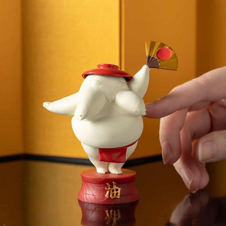 Peut inclure: Figurine blanche repr&eacute;sentant un lutteur de sumo avec un chapeau et un pagne rouges, tenant un petit &eacute;ventail. La figurine est sur une base rouge avec des caract&egrave;res japonais. L'&eacute;ventail est dor&eacute; avec un cercle rouge.