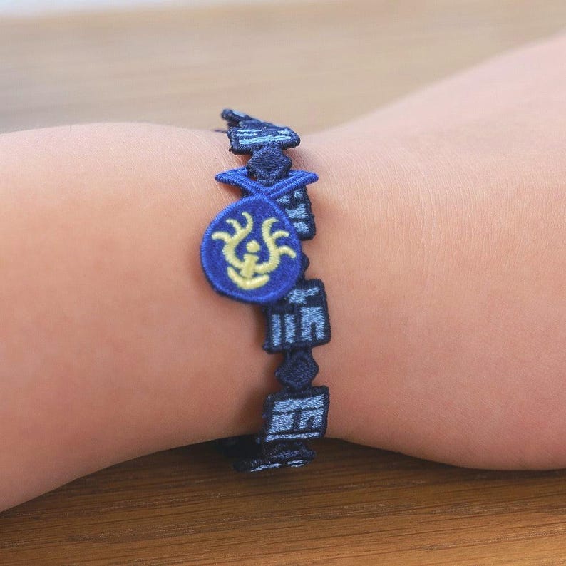 K&ouml;nnte beinhalten: Ein blaues, besticktes Armband mit einem sich wiederholenden geometrischen Muster und einem zentralen Anh&auml;nger. Der Anh&auml;nger ist ein blauer Kreis mit einem gelben Design. Das Armband wird am Handgelenk getragen.